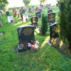 A cemetery with several headstones crafted by Memorial Masons, some adorned with flowers and wreaths, surrounded by green grass and trees on a sunny day.