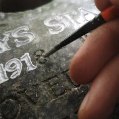 A hand uses a small paintbrush to carefully apply white paint to engraved letters and numbers on a dark stone surface, focusing on the number "1918," a delicate process often seen in Stone Restorations for headstones and cremation memorials.