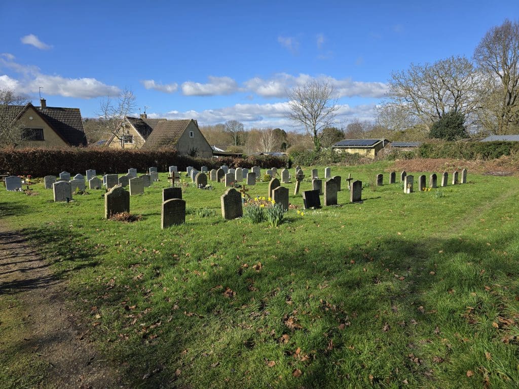 A peaceful cemetery with rows of headstones stands on green grass under a blue sky with scattered clouds. Leafless trees and a few houses are visible in the background, reflecting the work of skilled Memorial Masons.