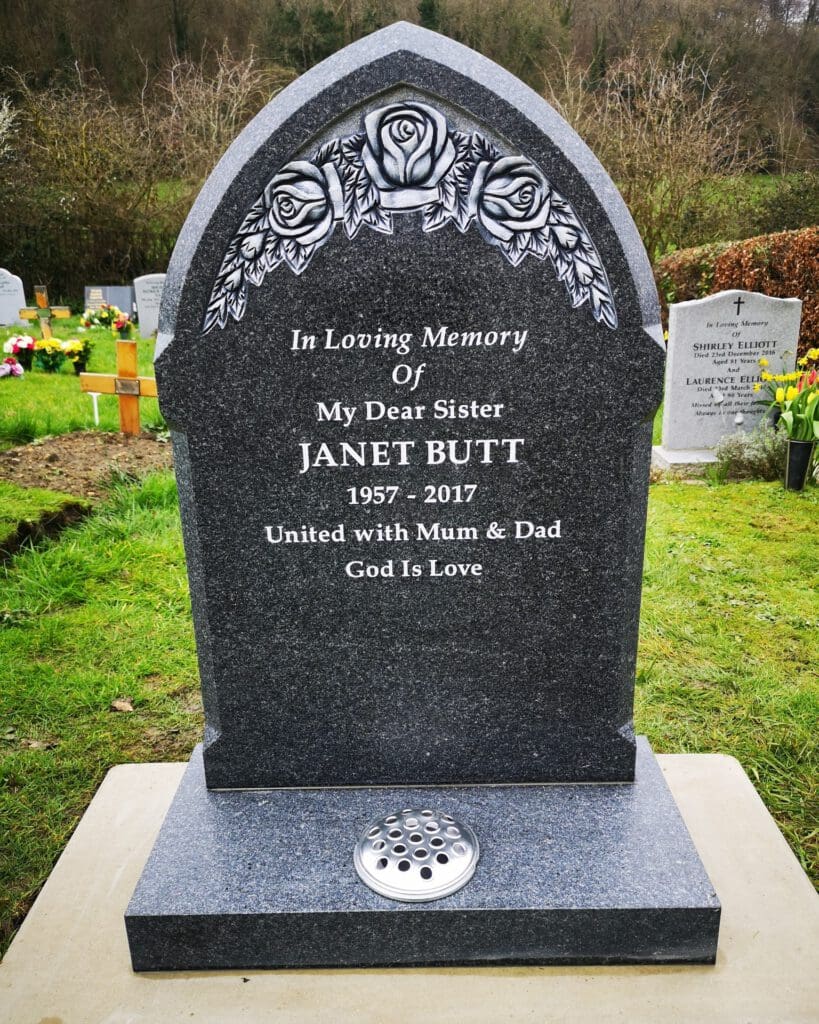 A dark tombstone engraved with roses at the top reads, "In Loving Memory of My Dear Sister Janet Butt 1957-2017. United with Mum & Dad. God Is Love." It stands on grass, surrounded by other headstones and flowers in the background.