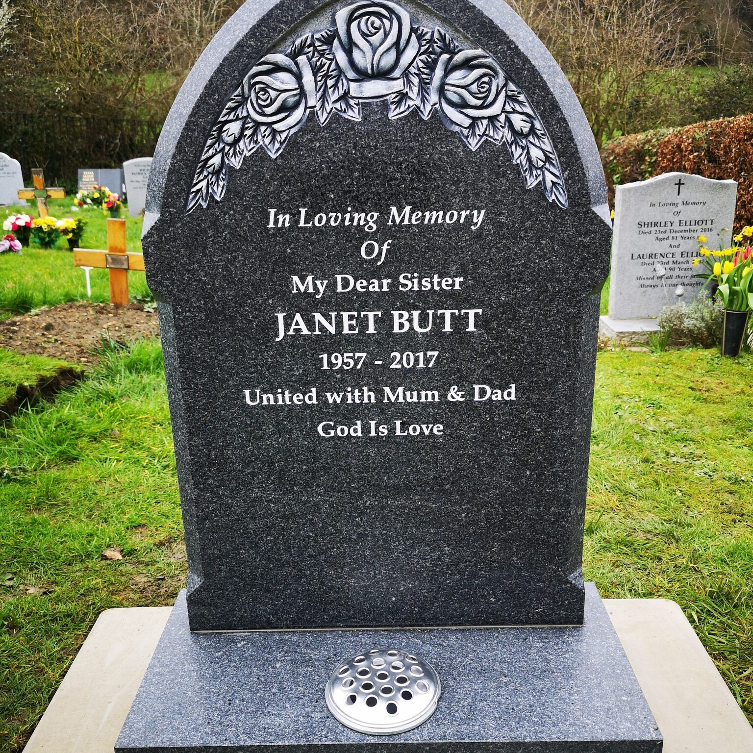 A dark tombstone engraved with roses at the top reads, "In Loving Memory of My Dear Sister Janet Butt 1957-2017. United with Mum & Dad. God Is Love." It stands on grass, surrounded by other headstones and flowers in the background.