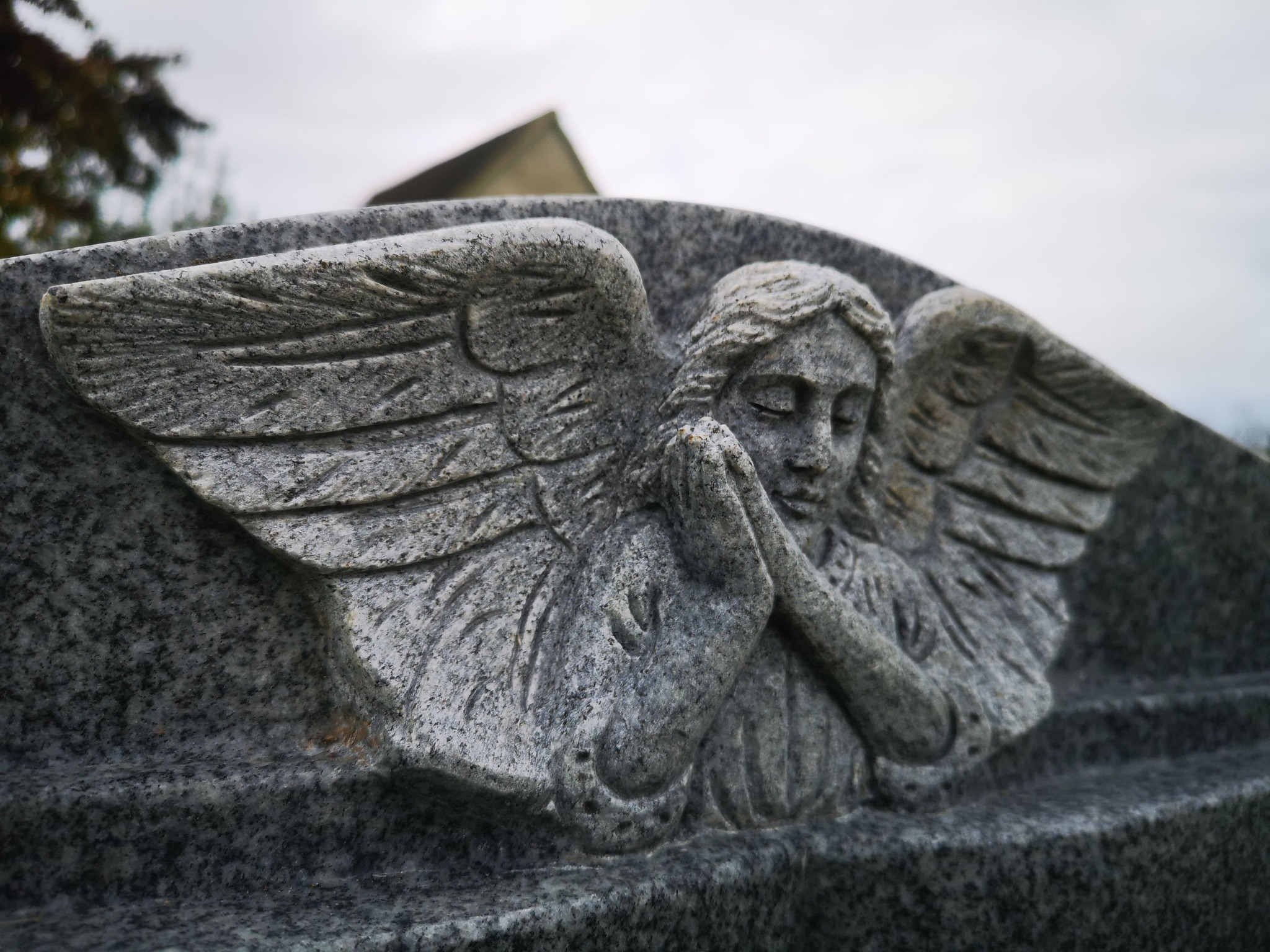 A close-up of a gray stone carving depicting an angel with wings folded behind, hands clasped in a praying position. The background is slightly blurred with hints of trees and a roof.