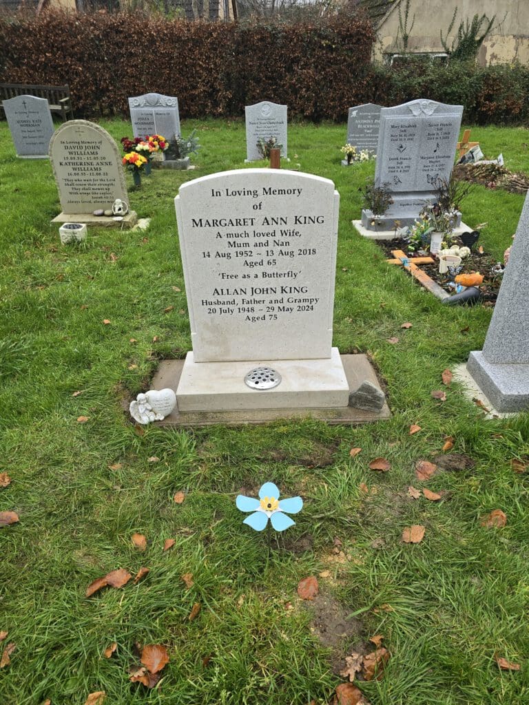 A cemetery scene with several headstones on grassy ground. The central headstone memorializes Margaret Ann King and Allan John King, adorned with a flower-patterned decoration and surrounded by other graves with flowers and vases.