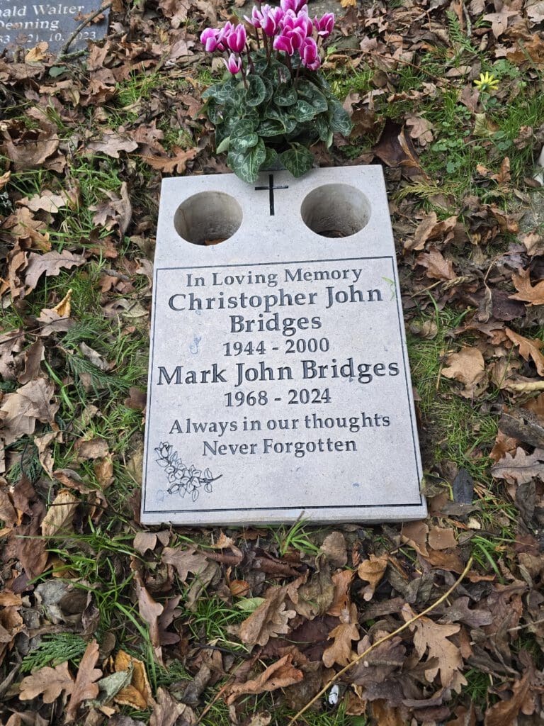 A gravestone with two flower holders shows inscriptions for Christopher John Bridges (1944-2000) and Mark John Bridges (1968-2024). It reads "Always in our thoughts Never Forgotten" with a small floral decoration. Pink flowers are on the stone.
