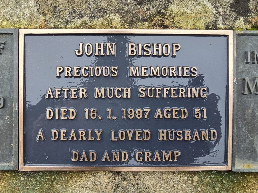 Plaque on a grave with the inscription: "John Bishop, precious memories after much suffering. Died 16.1.1987 aged 51. A dearly loved husband, dad and gramp." The background is weathered stone.