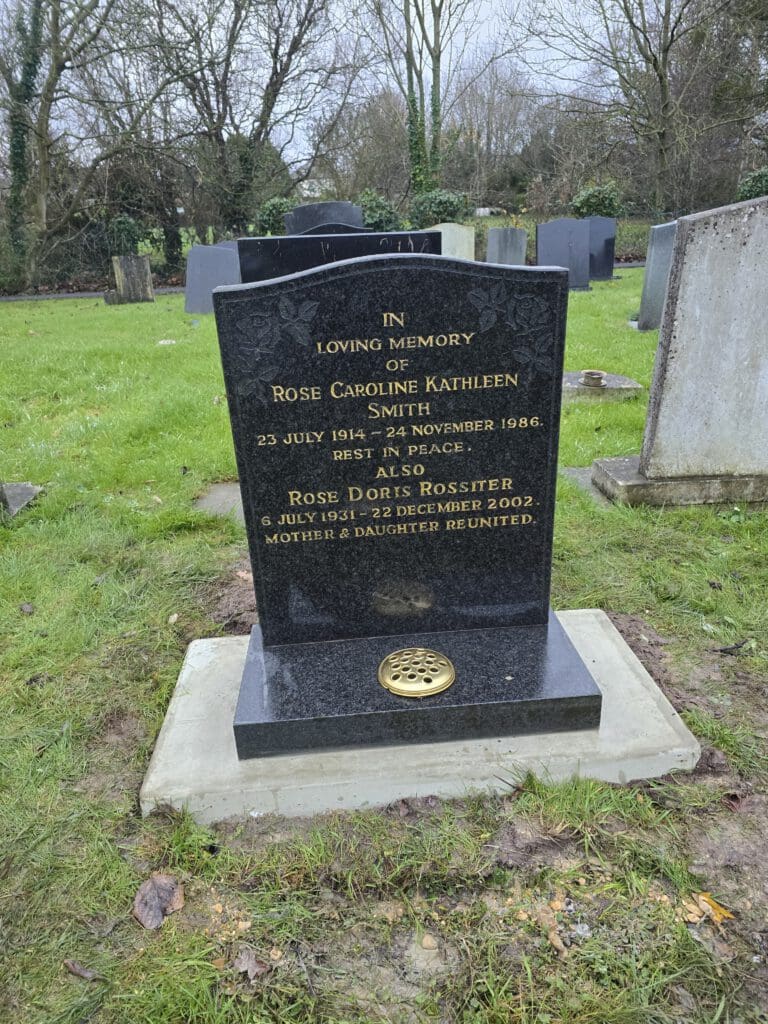 A black granite headstone in a cemetery with engraved text commemorating Rose Caroline Kathleen Smith and Rose Doris Rossiter. The grassy area is surrounded by other headstones and bare trees in the background.