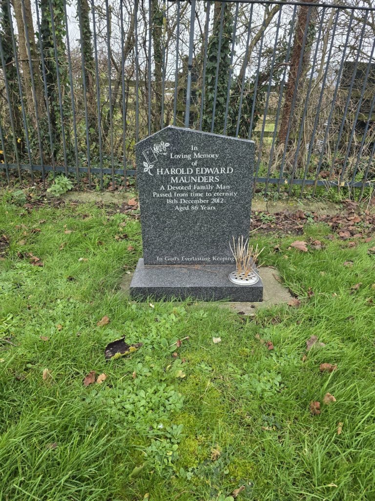 A gravestone surrounded by grass, dedicated to Harold Edward Maunders. The headstone has flowers and the inscription includes dates and a message of remembrance. A potted plant is placed in front of the stone. Trees and a fence are in the background.