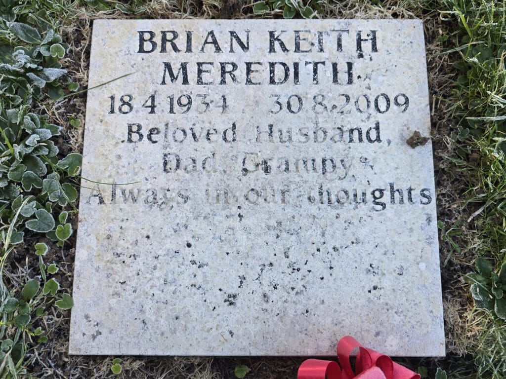 A memorial stone engraved with "Brian Keith Meredith, 18-4-1934 - 30-8-2009, Beloved Husband, Dad, Grampy. Always in our thoughts." It rests on grass with scattered clover leaves and a small red flower arrangement nearby.