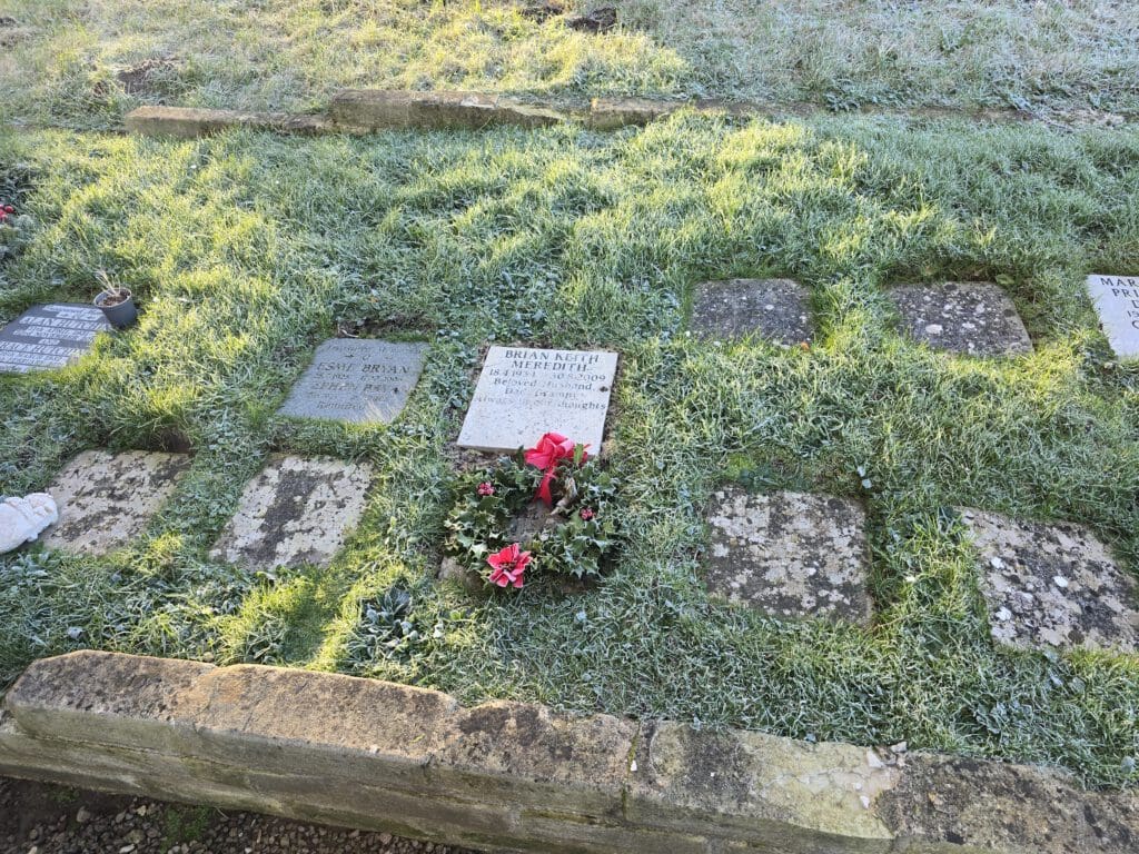 A grassy cemetery scene showing several worn and moss-covered headstones, some with unreadable text. A central headstone is decorated with a Christmas wreath featuring red bows and flowers. Frost covers the ground, suggesting a chilly day.