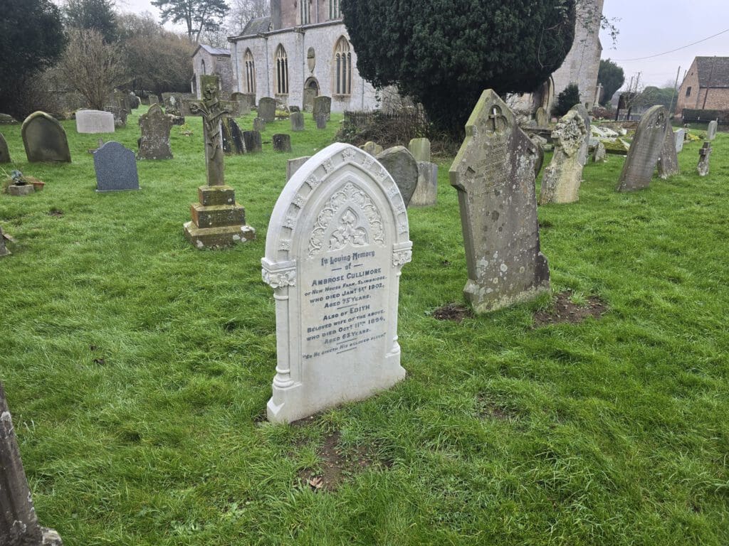 A historical cemetery with several weathered tombstones on green grass. One prominent white tombstone has visible engraved text. In the background, a large stone building, possibly a church, is partially visible with trees surrounding it.