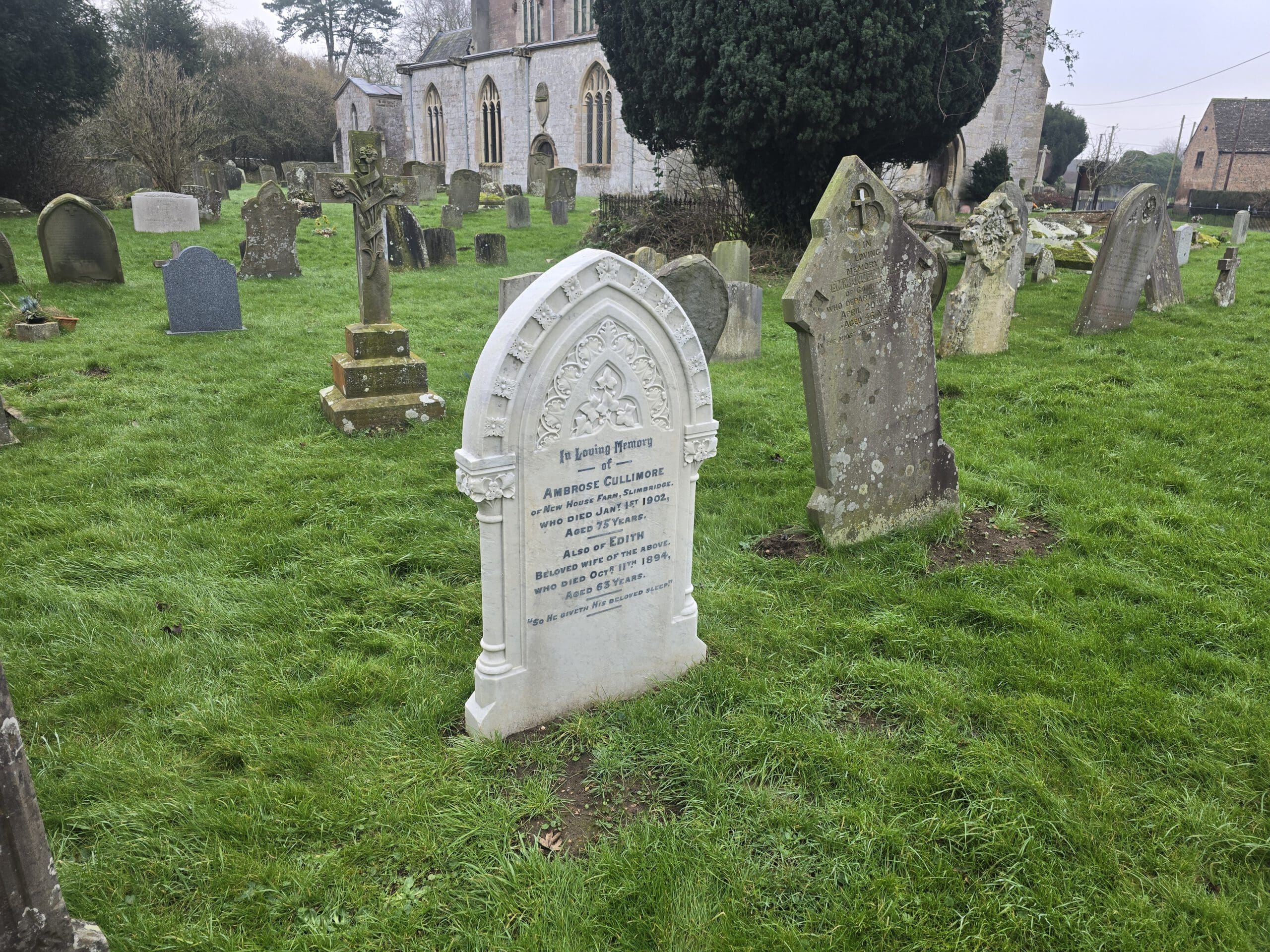 A historical cemetery with several weathered tombstones on green grass. One prominent white tombstone has visible engraved text. In the background, a large stone building, possibly a church, is partially visible with trees surrounding it.