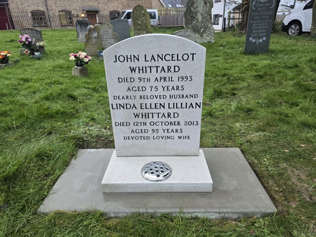A gravestone for John Lancelot Whittard, who died on 9th April 1993 at 75, and his wife Linda Ellen Lillian Whittard, who died on 12th October 2013 at 95. It’s set in a grassy cemetery with other tombstones and flowers in the background.