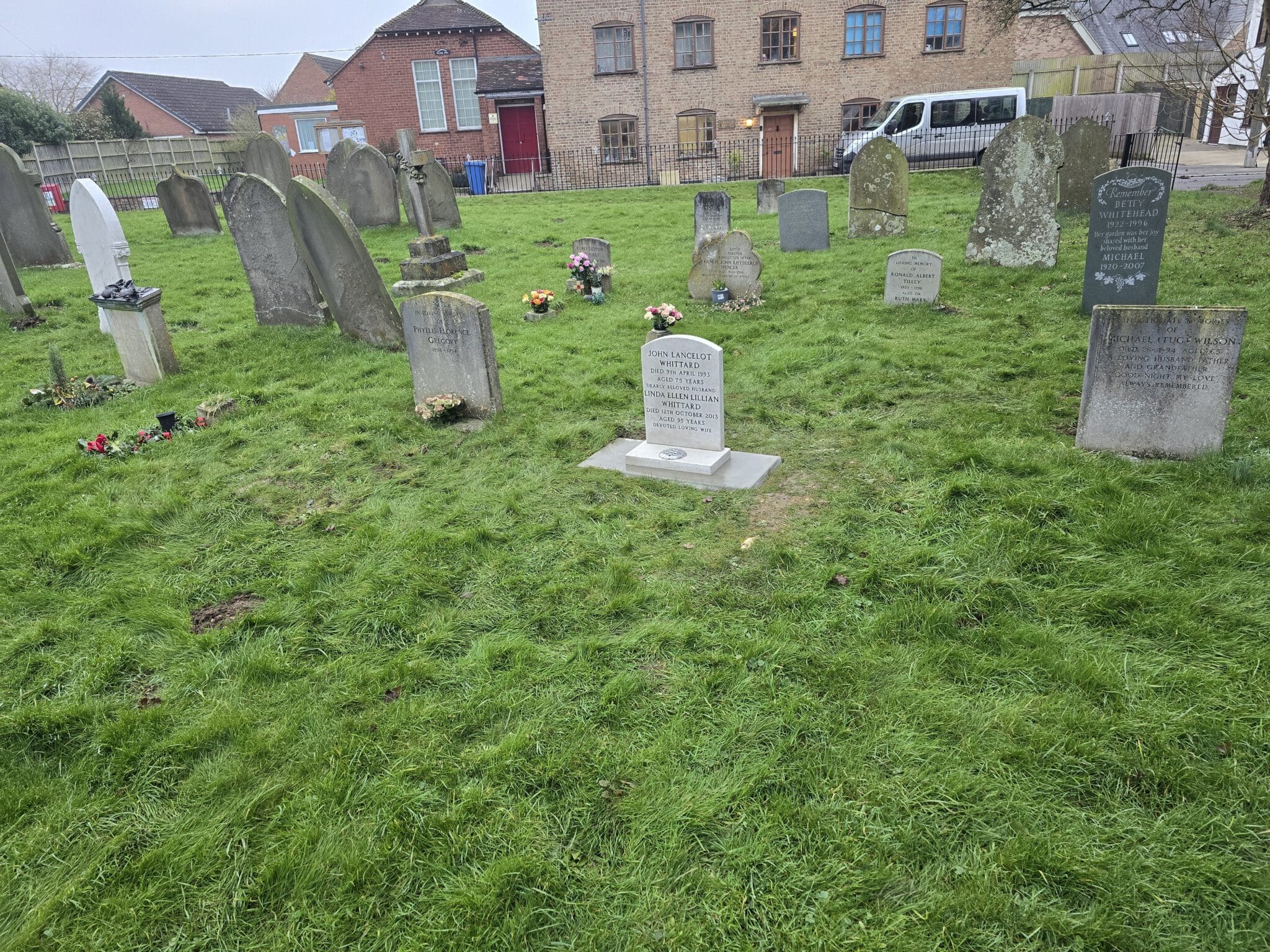 A cemetery with several weathered headstones on a grassy lawn. Flowers are placed by some graves. In the background, there are brick buildings and a white van parked near a fence. The sky appears overcast.