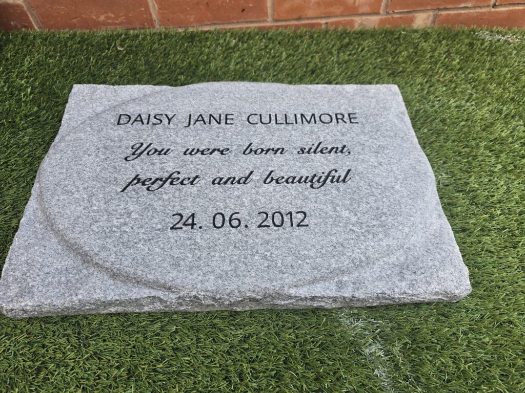 A rectangular gray stone memorial on grass, inscribed with: "DAISY JANE CULLIMORE. You were born silent, perfect and beautiful. 24.06.2012.