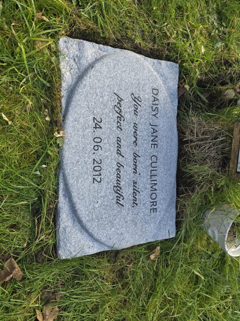 A small gray headstone on grass, engraved with "DAISY JANE CULLIMORE" and "You were born silent, perfect and beautiful" followed by the date "24.06.2012." A metal watering can is partially visible nearby.