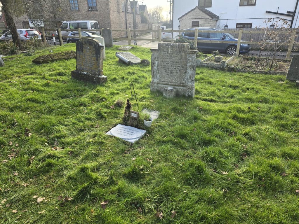 A grassy cemetery with old, weathered gravestones, including one close to the foreground. The background shows a road and parked cars, with brick and white buildings partially visible. Sparse trees and a wooden fence border the area.