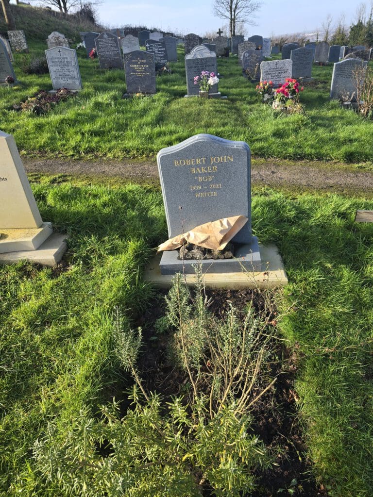 A headstone in a cemetery reads "Robert John Baker, 1940-2004, Writer." The grave has a bouquet of dried flowers placed on it. Green grass and other headstones surround the area under a clear sky.