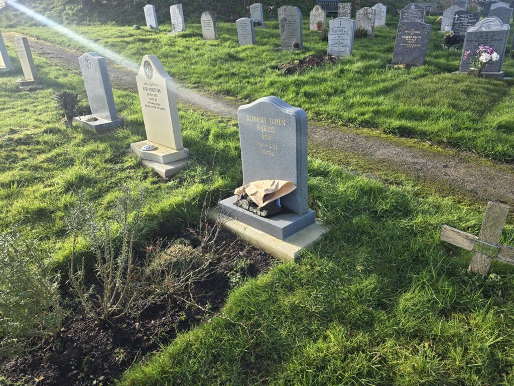 A cemetery with several headstones on a grassy area. One prominent gray headstone has a paper bag and stones placed on its base. A small path runs between the rows of graves, and the sun casts long shadows.