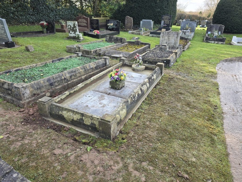 A cemetery scene with several gravestones and flower arrangements on various graves. The grass is green and well-maintained, and the sky is overcast, creating a serene atmosphere. Some graves have rectangular stone borders.