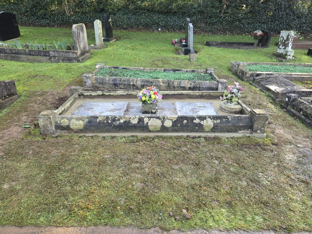 A weathered gravestone in a cemetery with a stone border, featuring a vase of colorful flowers at its center. Surrounding graves and a lush green hedge are visible in the background.