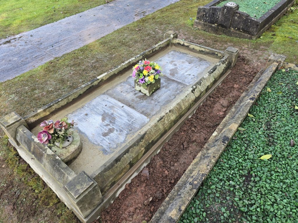 A weathered cemetery plot with a stone grave marker. Colorful flowers in a planter sit atop the grave, flanked by additional petals. The grass surrounding the grave is trimmed, and nearby paths are visible.