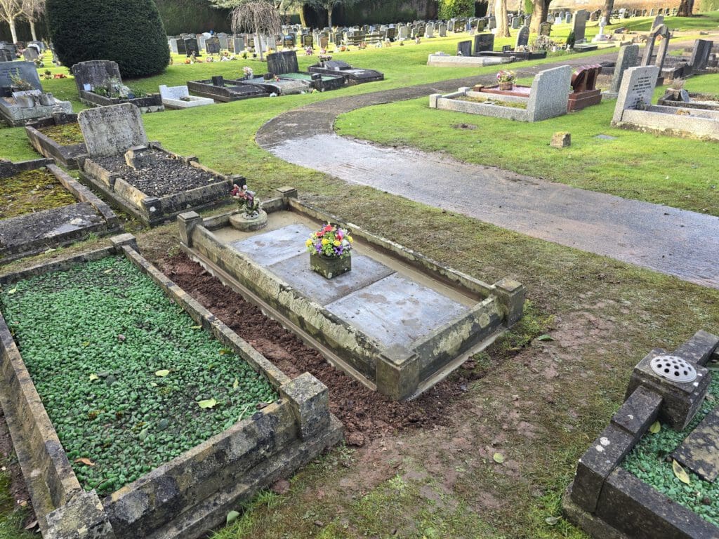 A peaceful cemetery scene with rows of gravestones and grassy paths. A prominent grave in the foreground is adorned with floral arrangements. A curved pathway winds through the well-kept grounds, bordered by lush greenery and trees.