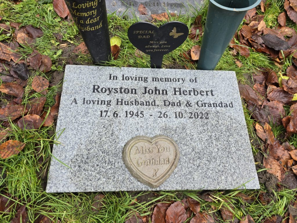 A gravestone with the inscription "In loving memory of Royston John Herbert, a loving Husband, Dad & Grandad, 17.6.1945 - 26.10.2022." It is surrounded by fallen leaves and a small heart-shaped plaque with "Miss You Granddad" on it.