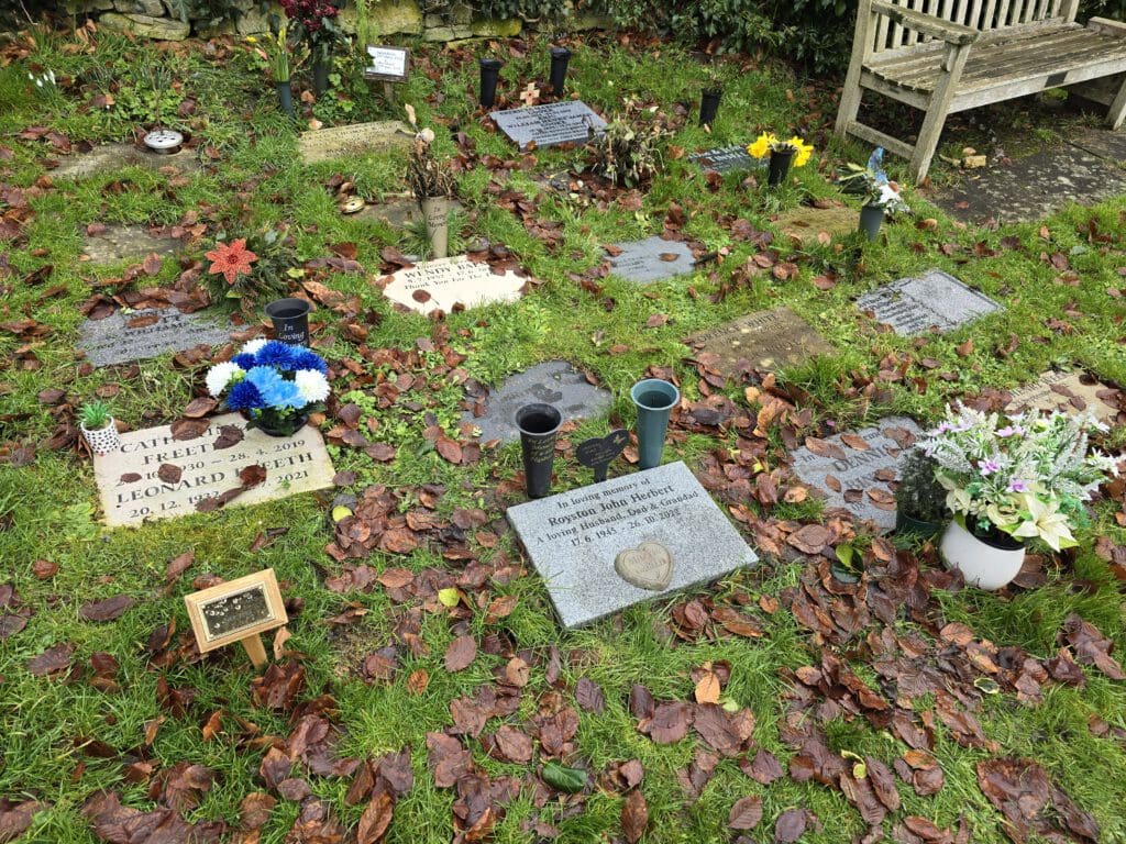 A cemetery scene with multiple flat gravestones on grassy ground, covered in autumn leaves. Some headstones have flowers and small vases. A wooden bench is visible in the background.