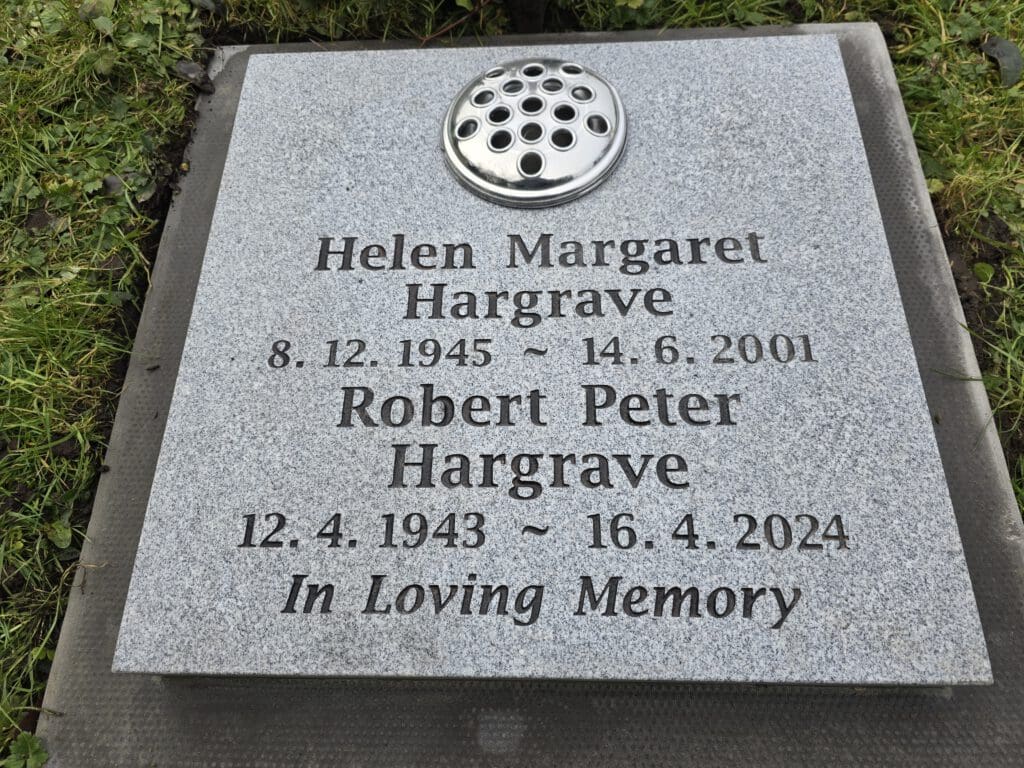 A gray granite headstone with engraved names: Helen Margaret Hargrave (1945-2001) and Robert Peter Hargrave (1943-2024). It reads "In Loving Memory" and features a vase holder with holes for flowers.