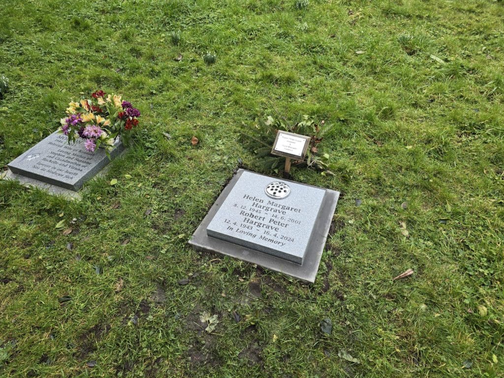 A grassy area with two memorial plaques on the ground. One plaque is adorned with a bouquet of colorful flowers, while the other is a light gray stone with engraved text and a circular emblem above it.