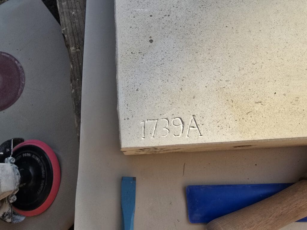 Close-up of a stone slab with the engraving "1739A" in the corner. A blue flat tool and a rounded tool with a handle are nearby on a beige surface.