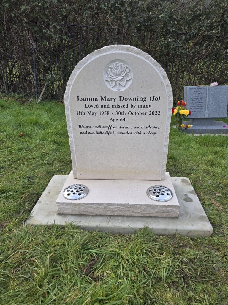 A gravestone with an engraved rose at the top, commemorating Joanna Mary Downing (Jo), who lived from May 11, 1958, to October 30, 2022. It reads, "Loved and missed by many." There's a quote about life and dreams. Flowers are nearby.