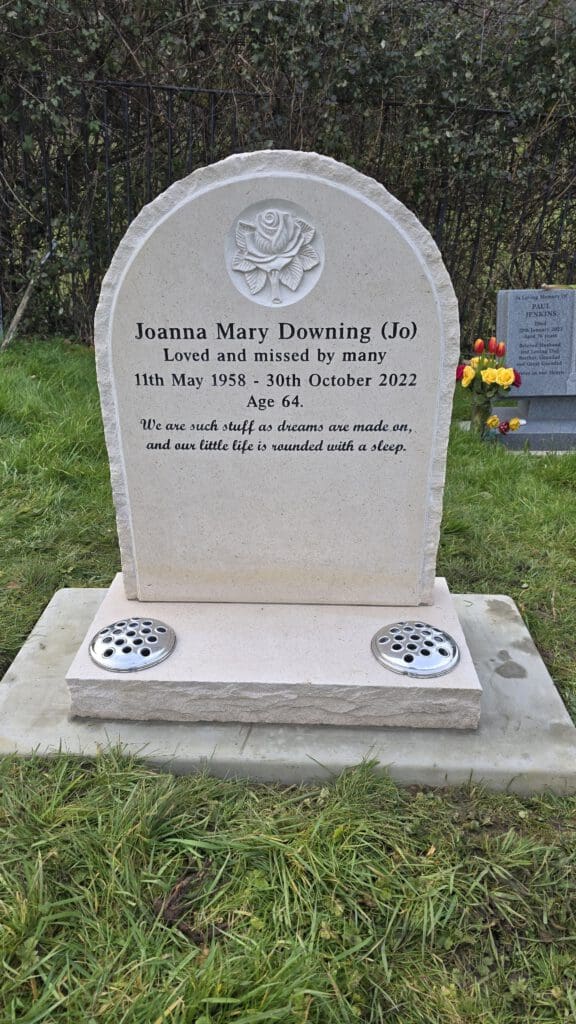 A gravestone with an engraved rose at the top, commemorating Joanna Mary Downing (Jo), who lived from May 11, 1958, to October 30, 2022. It reads, "Loved and missed by many." There's a quote about life and dreams. Flowers are nearby.