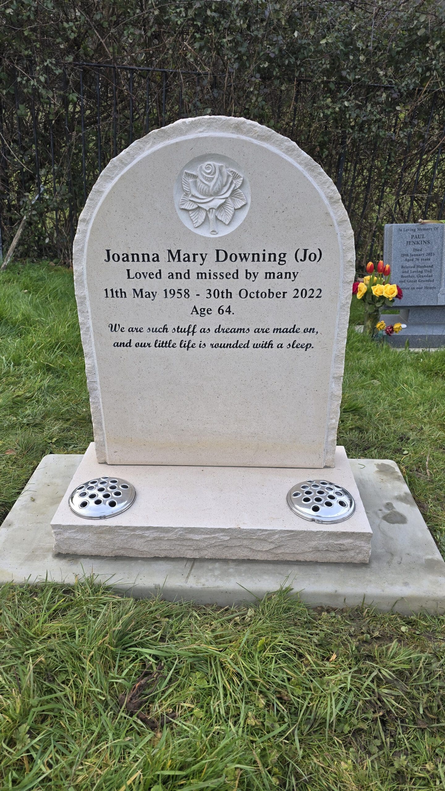 A gravestone with an engraved rose at the top, commemorating Joanna Mary Downing (Jo), who lived from May 11, 1958, to October 30, 2022. It reads, "Loved and missed by many." There's a quote about life and dreams. Flowers are nearby.