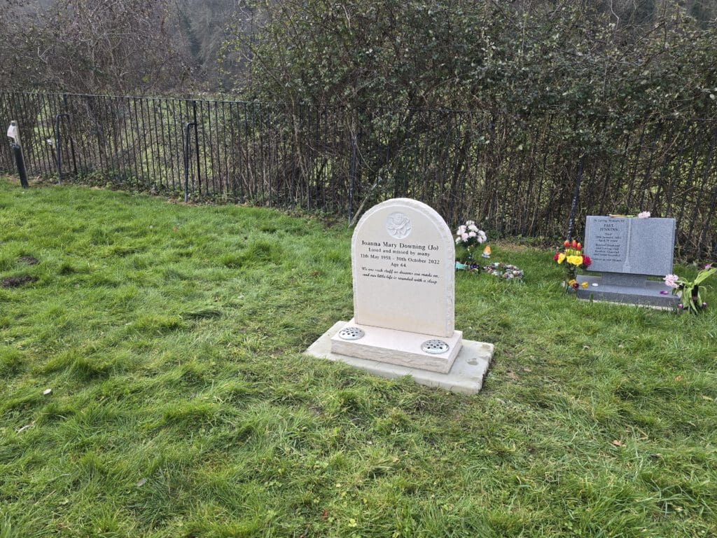 A white gravestone on grassy ground, with some text visible. Nearby, a smaller stone is adorned with colorful flower arrangements. A metal fence and trees are in the background.