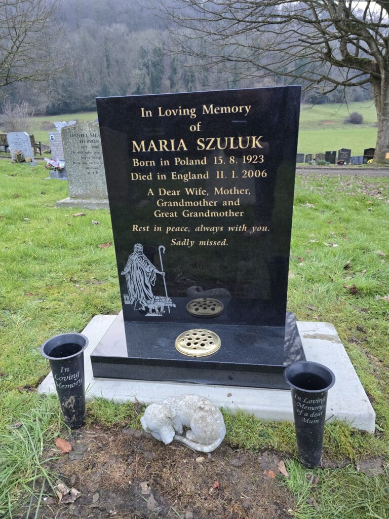 A black headstone in a cemetery with trees and rolling hills in the background. It reads "In Loving Memory of Maria Szuluk" with her birth and death dates, followed by a dedication. Two black vases with inscriptions are placed on either side.