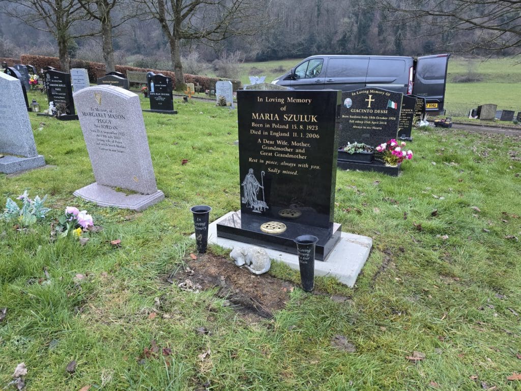 A black gravestone in a cemetery, inscribed with the name Maria Szulik, with candles and a small white statue at its base. The grave is set in a grassy area with other tombstones nearby. A black van is parked in the background, trees in view.