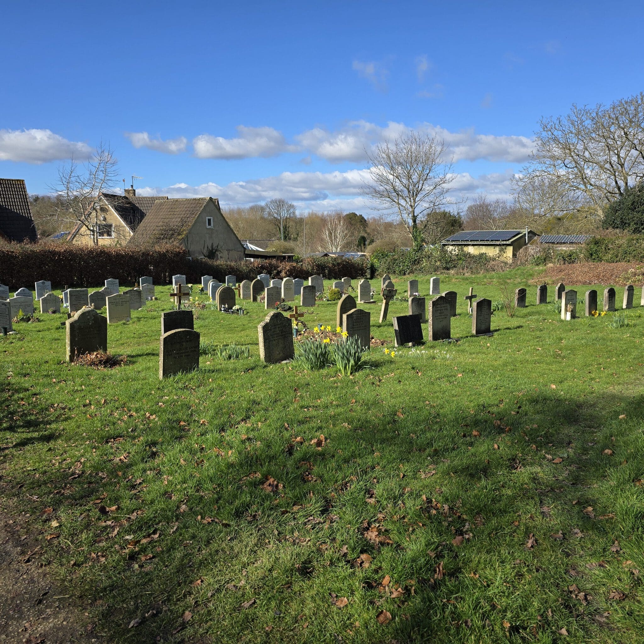Sunny graveyard scene with rows of headstones on green grass. A quaint stone building is visible in the background, surrounded by leafless trees and shrubs. The sky is blue with scattered clouds.