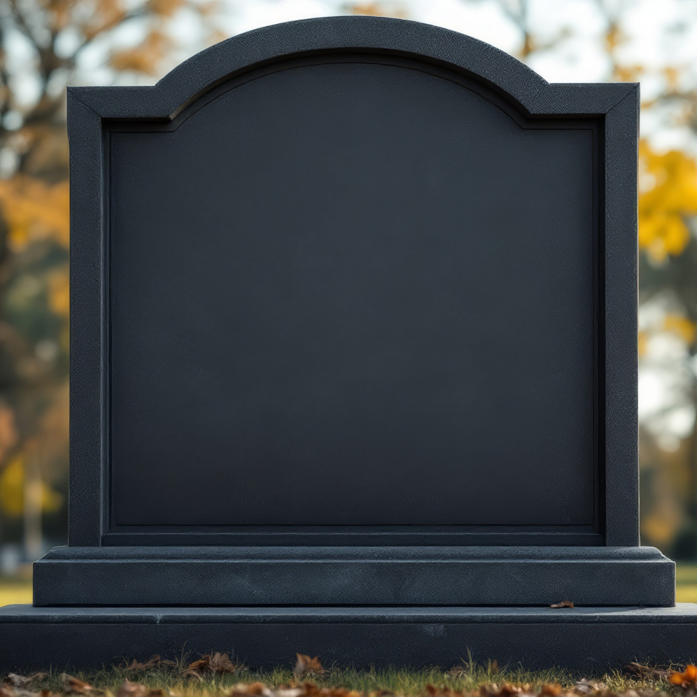 A blank, arched headstone stands on a grassy area adorned with fallen autumn leaves. Trees with yellow and orange foliage are blurred in the background, suggesting an autumn setting.
