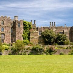 A large, historic stone castle with multiple chimneys and turrets, surrounded by green lawns and flowering shrubs under a blue sky—skilled memorial masons once crafted cremation memorials for those resting nearby.