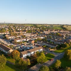 Aerial view of a suburban neighborhood with rows of houses, surrounded by greenery and fields. Two hot air balloons are visible in the distant sky. The sun casts a warm light, creating a peaceful atmosphere.