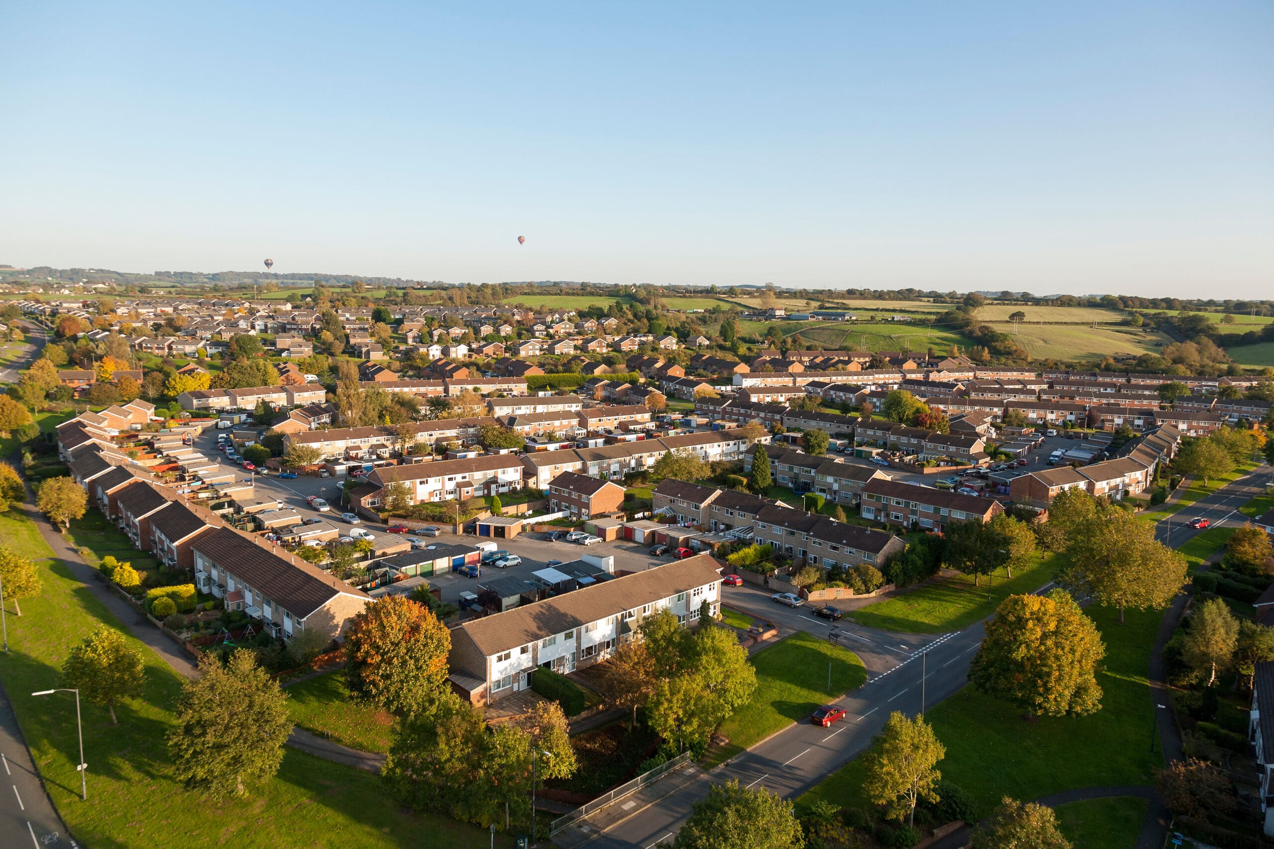 Aerial view of a suburban neighborhood with rows of houses, surrounded by greenery and fields. Two hot air balloons are visible in the distant sky. The sun casts a warm light, creating a peaceful atmosphere.