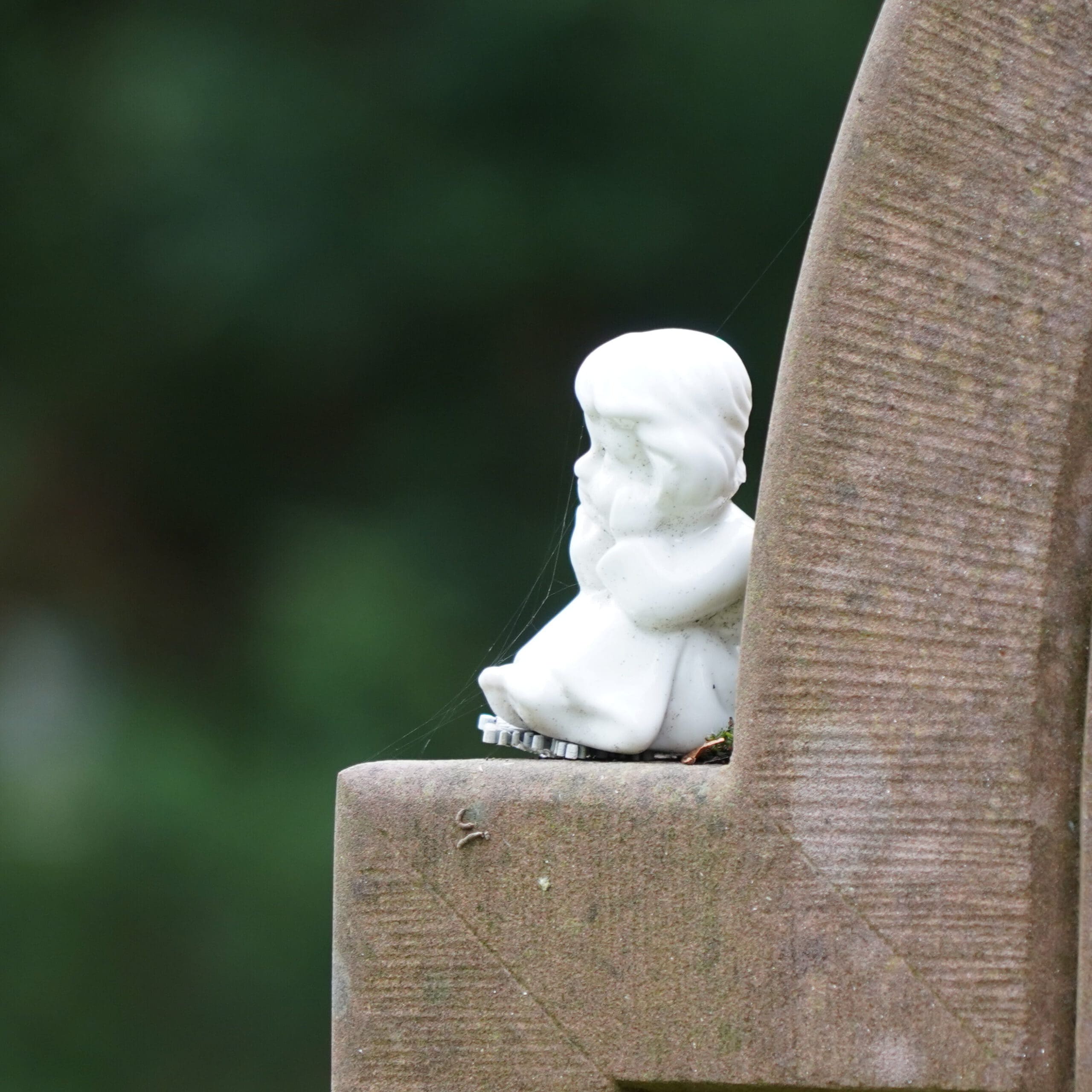 A small white statue of a child sitting and resting its chin in its hands is placed on the edge of a stone monument. The background is blurred greenery, emphasizing the contemplative pose of the figure.