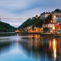 A suspension bridge at dusk spans across a river, with reflections of city lights, colorful buildings, and green hills—its stonework reminiscent of expert Memorial Masons and Stone Restorations—mirrored beautifully on the water below.