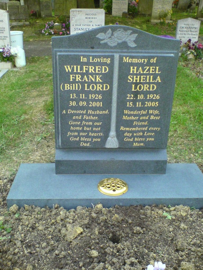 A headstone for Wilfred Frank (Bill) Lord and Hazel Sheila Lord. Wilfred's dates: 13.11.1926 - 30.09.2001, Hazel's dates: 22.10.1926 - 15.11.2005. Tribute mentions family roles and love. Surrounding graves and flowers are visible in the background.