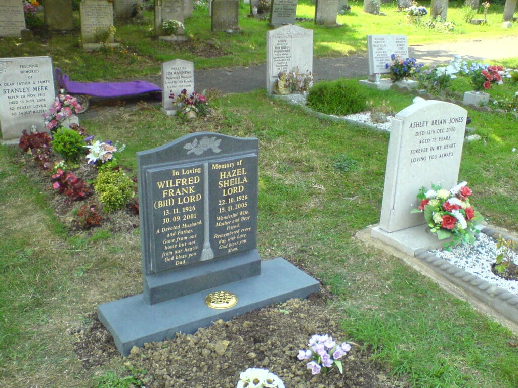 A cemetery scene with two gravestones. The left gravestone is for Wilfred Frank Lord and Hazel Sheila Lord, flanked by flowers. The right gravestone is for Ashley Brian Jones, surrounded by floral arrangements and small plants. Other gravestones in the background.