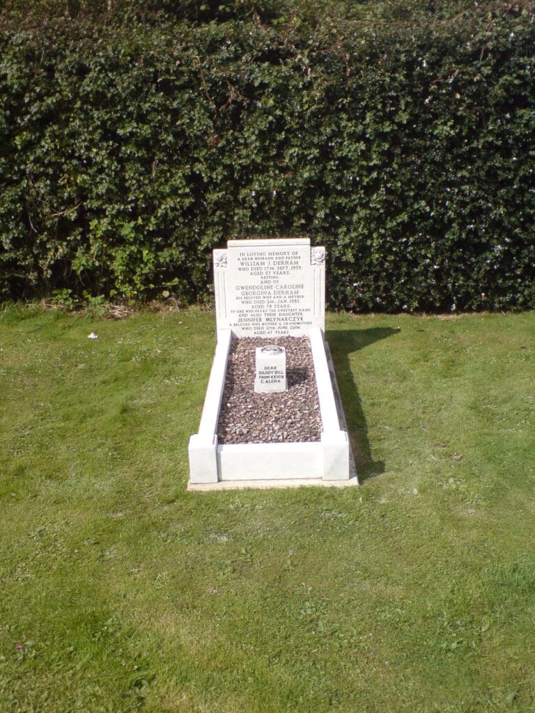 A white gravestone with inscriptions is set on a rectangular plot filled with small stones. The plot is surrounded by a well-maintained grassy area, and a tall, dense hedge is visible in the background.
