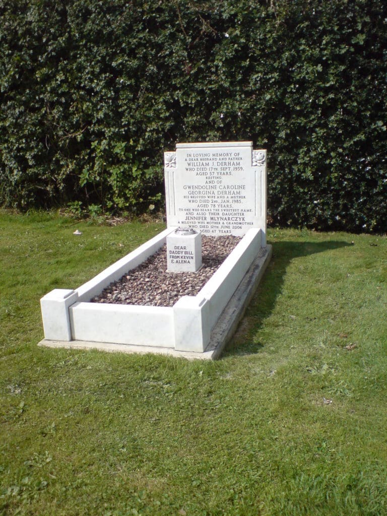A white marble gravestone surrounded by grass, with a small plaque in front, marking the resting place of multiple individuals. The headstone is inscribed with names and dates, set against a backdrop of green foliage.