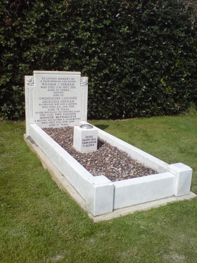 A gravestone set on a grass lawn, framed by a hedge. It features engraved names and dates, with a smaller plaque on top of a gravel area. The stone is white and rectangular, contrasting with the dark pebbles and green background.