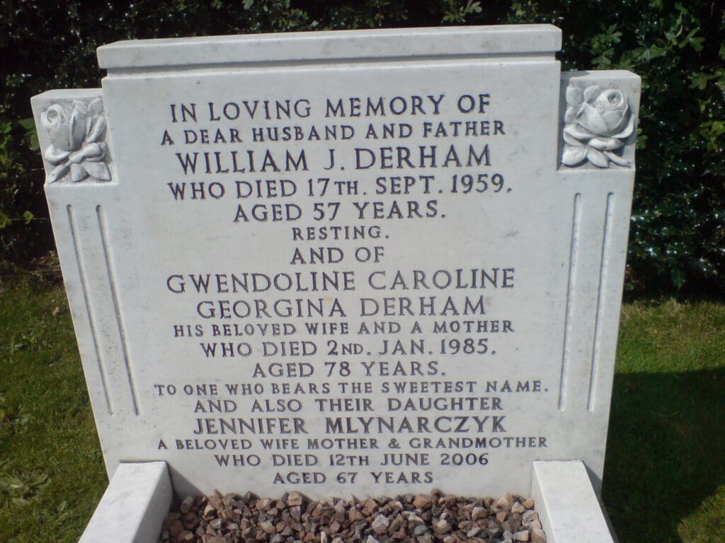 A white gravestone with inscriptions in memory of William J. Derham, who died in 1959, aged 57; Gwendoline Caroline Georgina Derham, who died in 1985, aged 78; and Jennifer Mlynarczyk, who died in 2006, aged 67. Floral carvings adorn the stone.